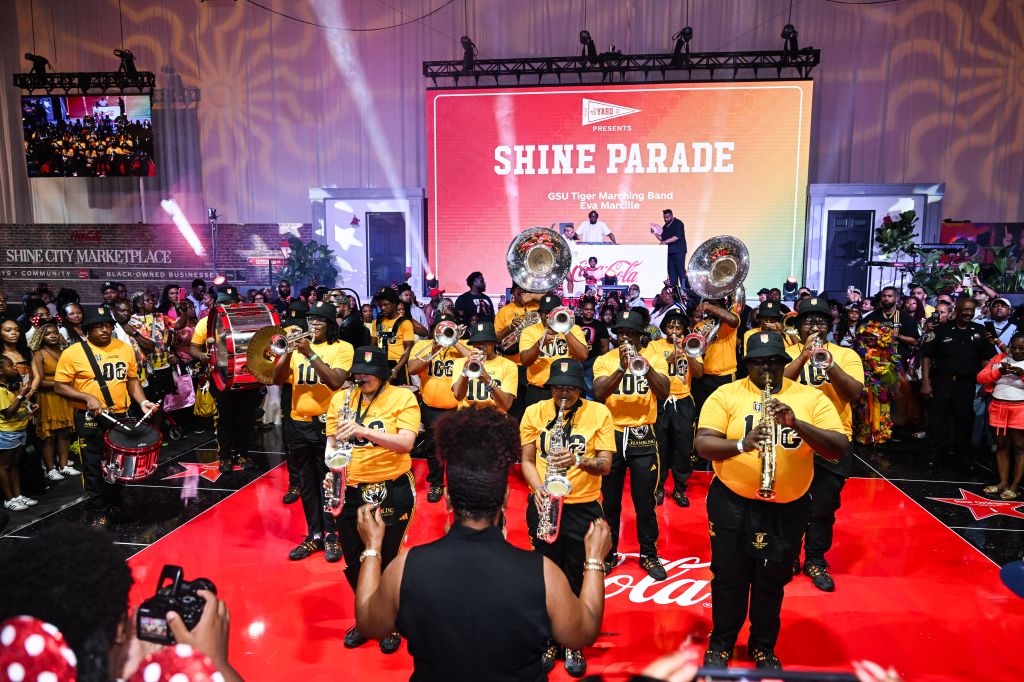 A large marching band in yellow uniforms performing on a stage with a "Shine Parade" backdrop and a red carpet.