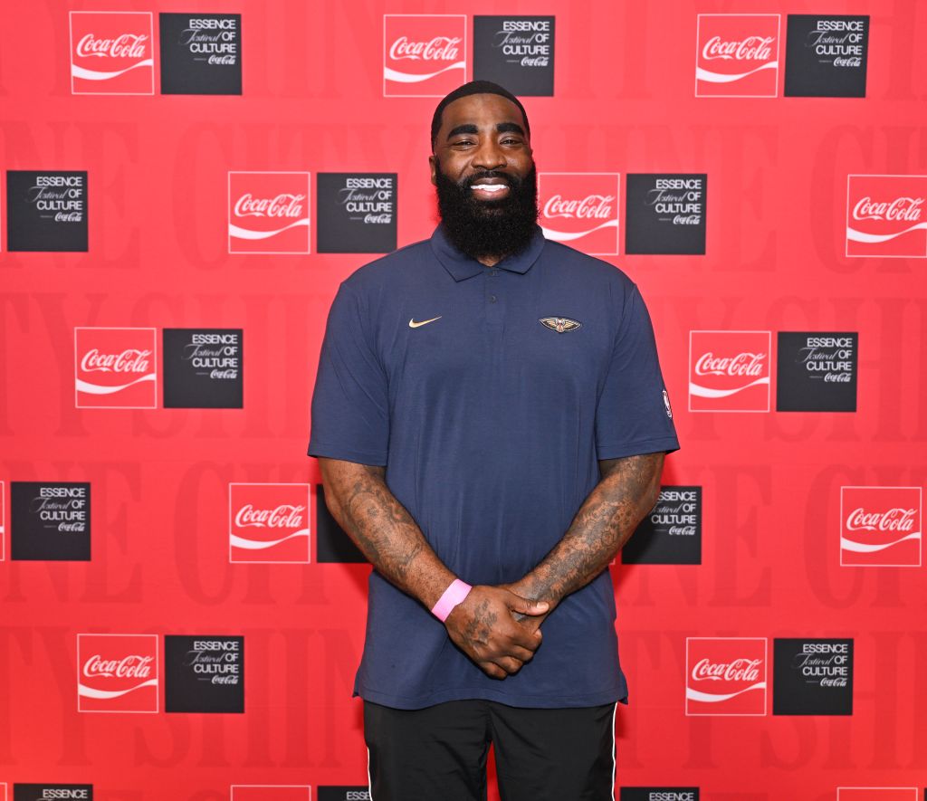A bearded man wearing a navy blue Nike polo shirt stands in front of a red and black Coca-Cola "Essence of Culture" backdrop.