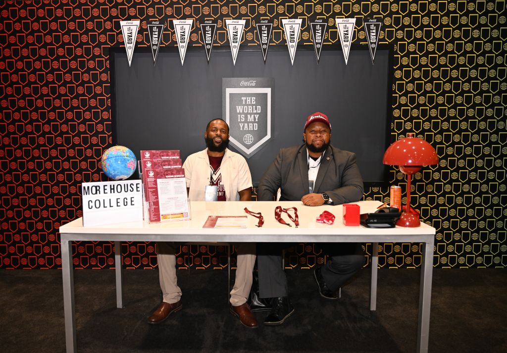 Two men sitting at a table with Coca-Cola branding, "The world is my yard" banner, and "More House College" signage.