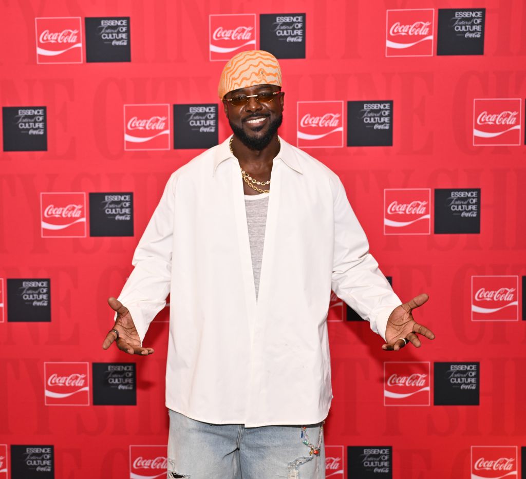 A smiling man wearing a white coat and a patterned hat stands in front of a red background with Coca-Cola and "Essence of Culture" logos.