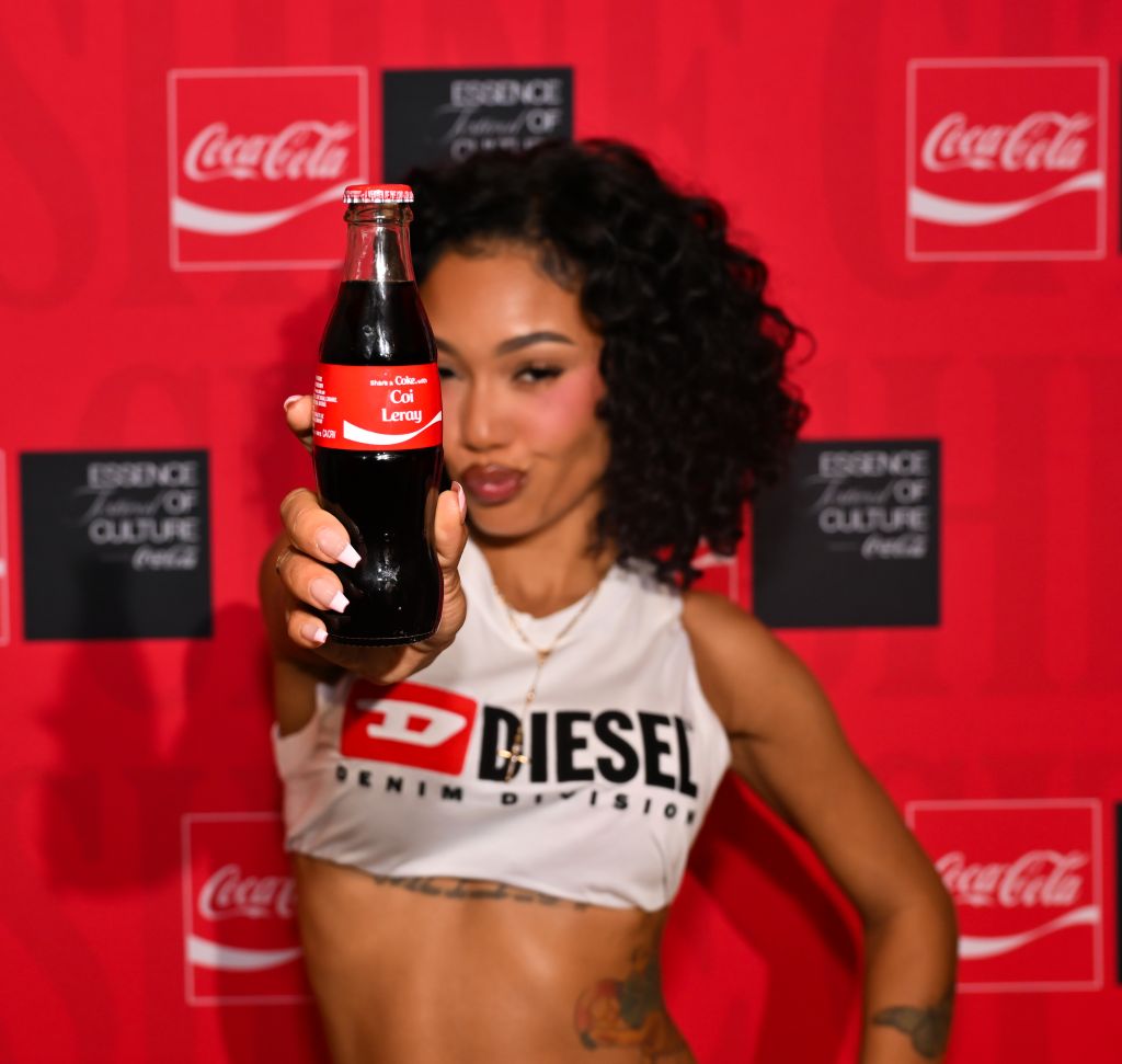 A woman holding a Coca-Cola bottle and wearing a Diesel brand crop top stands in front of Coca-Cola branding.