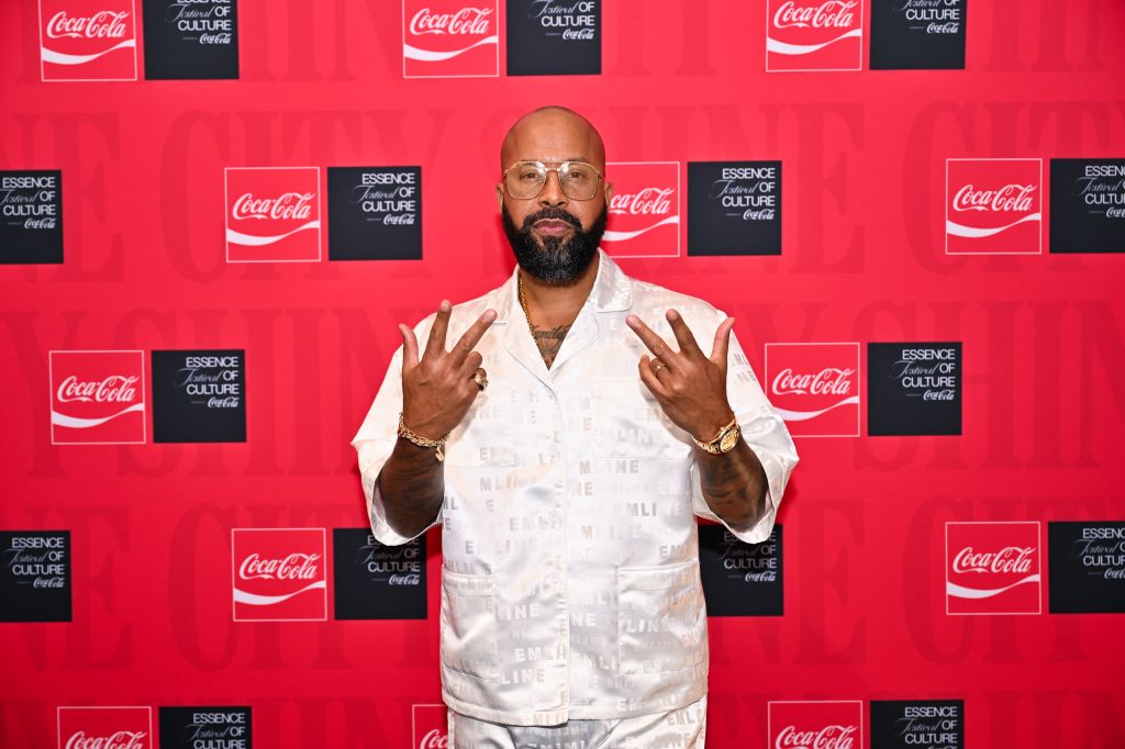 A man with a beard and glasses wearing a white suit with Coca-Cola branding, making a hand gesture in front of a red and black Coca-Cola backdrop.