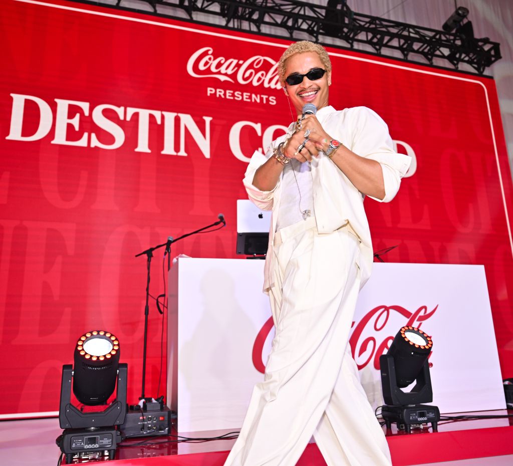 A performer in a white suit standing on a stage with a large Coca-Cola logo and the text "Destin Cole" displayed behind them.