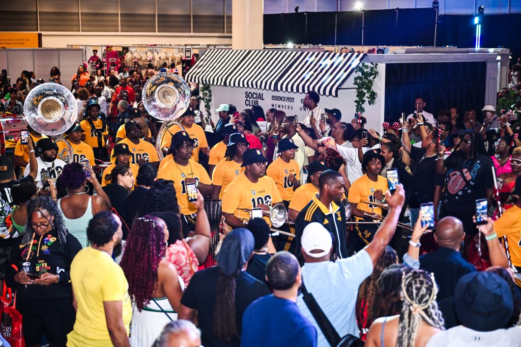 A crowded nighttime scene with a large group of people wearing yellow shirts, some holding brass instruments, gathered around a striped canopy with "Social Club" signage.