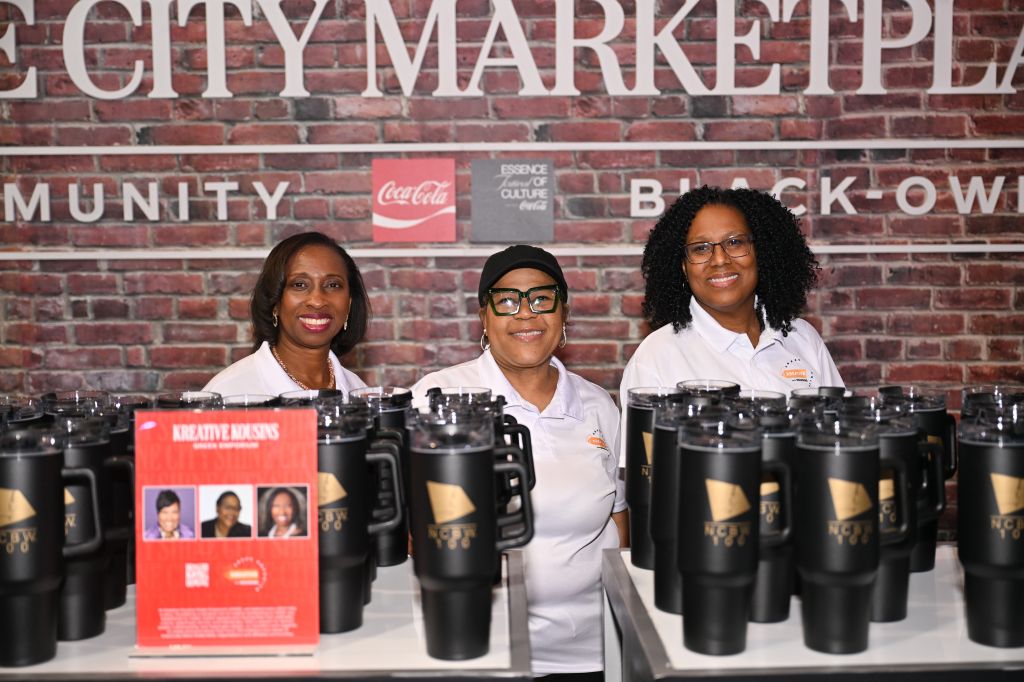 Three smiling African American women in white uniforms stand behind a display of Coca-Cola branded cups and mugs in a City Market retail setting.