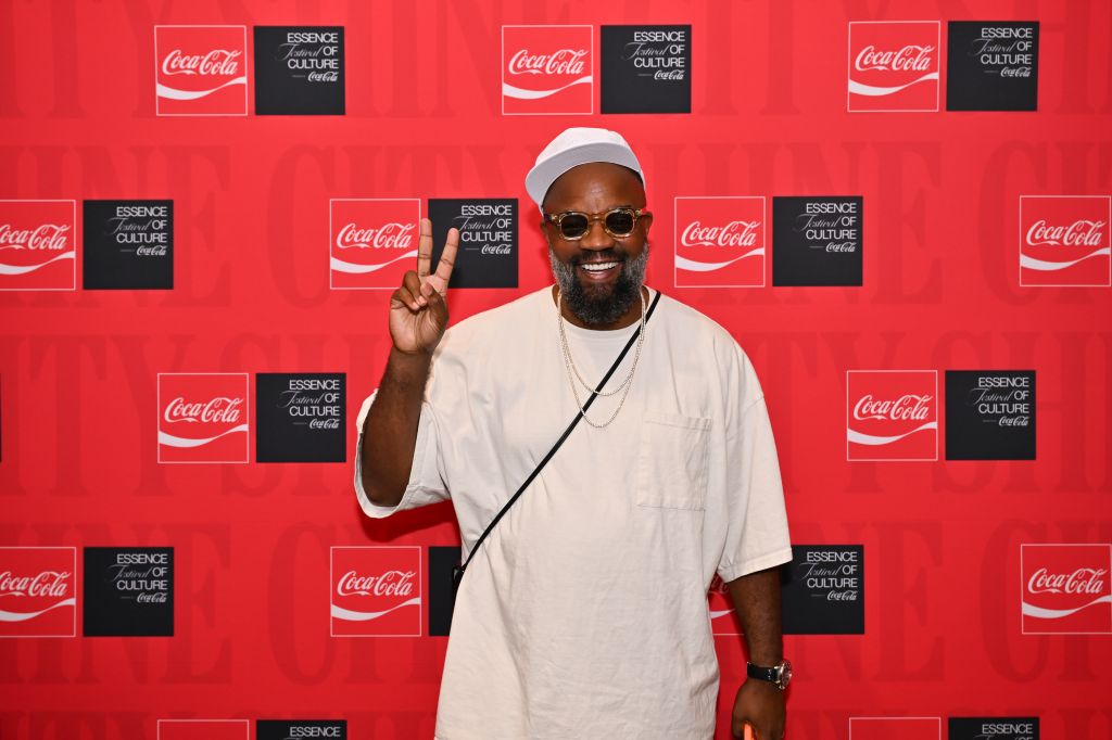 A smiling man with a beard wearing sunglasses and a white shirt, making a peace sign in front of a red background with Coca-Cola and "Essence of Culture" branding.