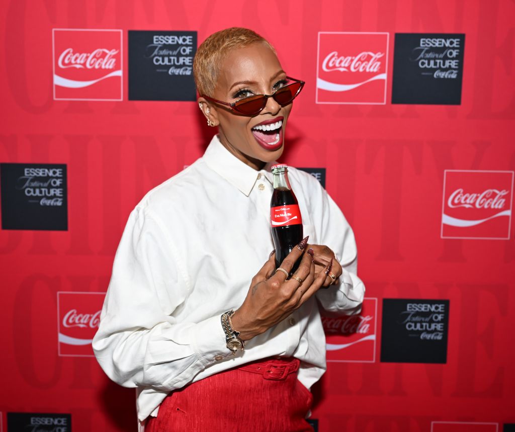 A smiling woman in a white shirt holding a Coca-Cola bottle, standing in front of Coca-Cola and "Essence of Culture" branding.