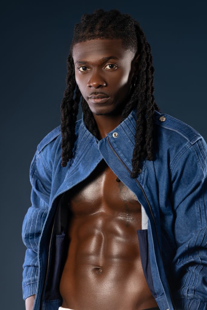 A shirtless Black man with dreadlocks wearing a denim jacket stands against a dark background, displaying a muscular physique.