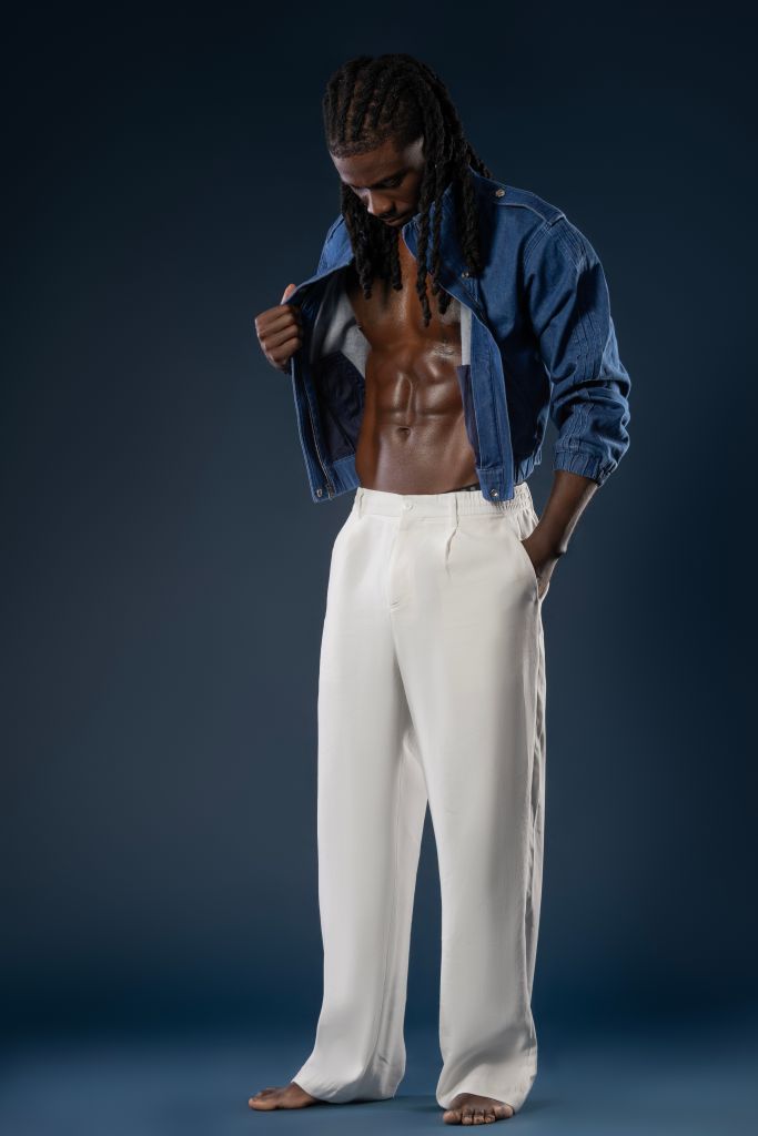 A shirtless man with dreadlocks wearing a denim jacket, standing against a dark background.