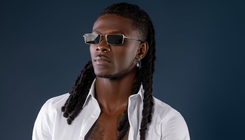 A shirtless Black man with dreadlocks wearing sunglasses and a white shirt, standing against a dark background.