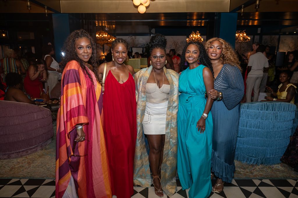 A group of five stylishly dressed Black women posing together in an elegant, dimly lit setting with chandeliers and patterned floors.