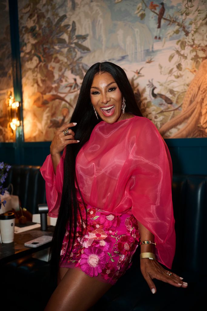 A smiling woman in a bright pink dress and cape stands in front of a colorful mural backdrop, gesturing with her hands.