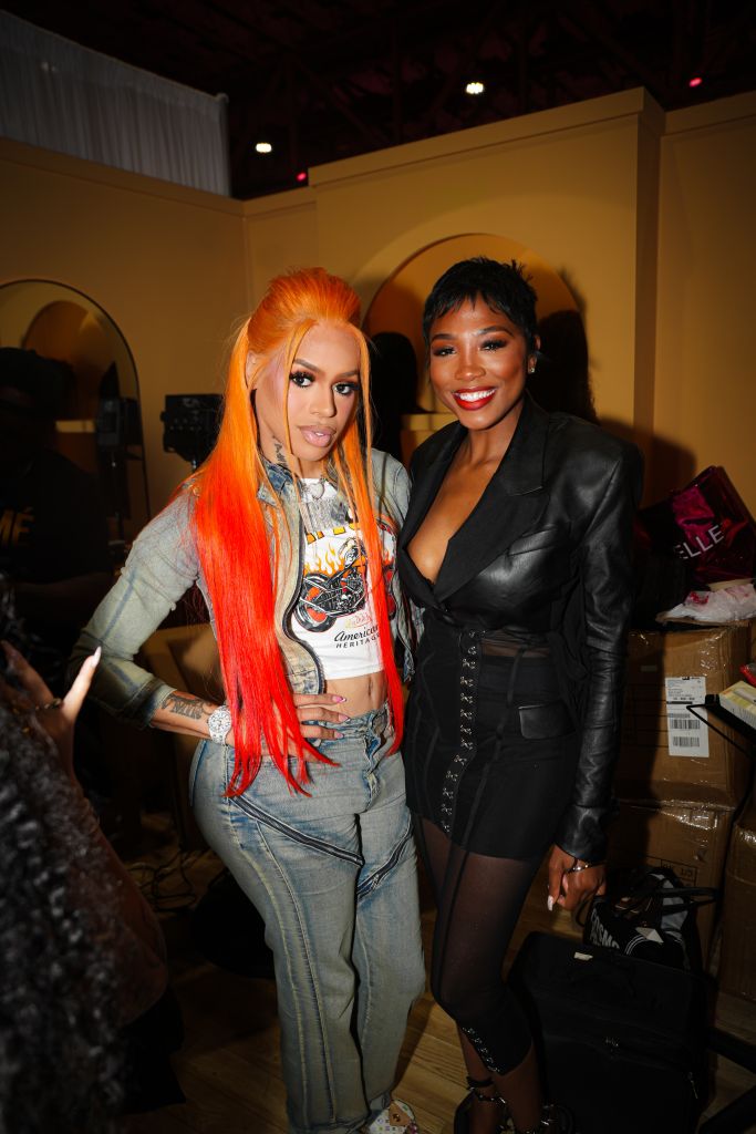 Two women posing together, one with long orange hair wearing a graphic t-shirt and the other in a black leather jacket and shorts.