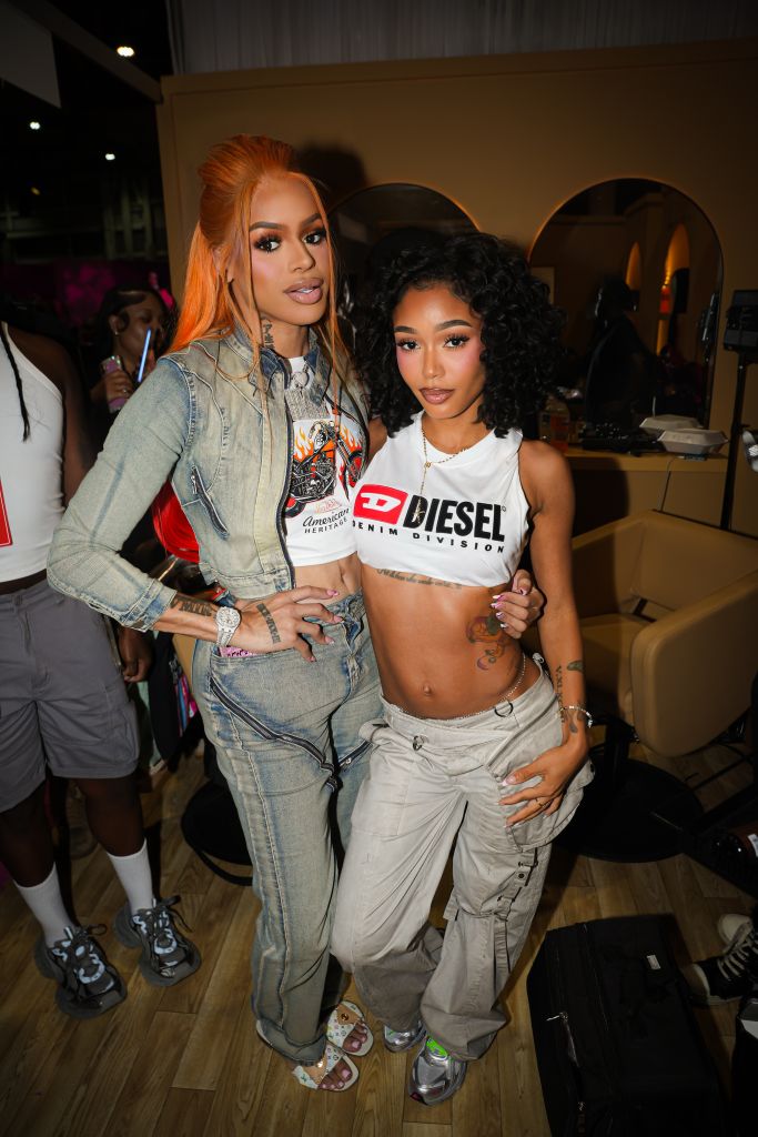 Two young women posing together, one with orange hair wearing a denim jacket and the other with curly dark hair wearing a white Diesel crop top.