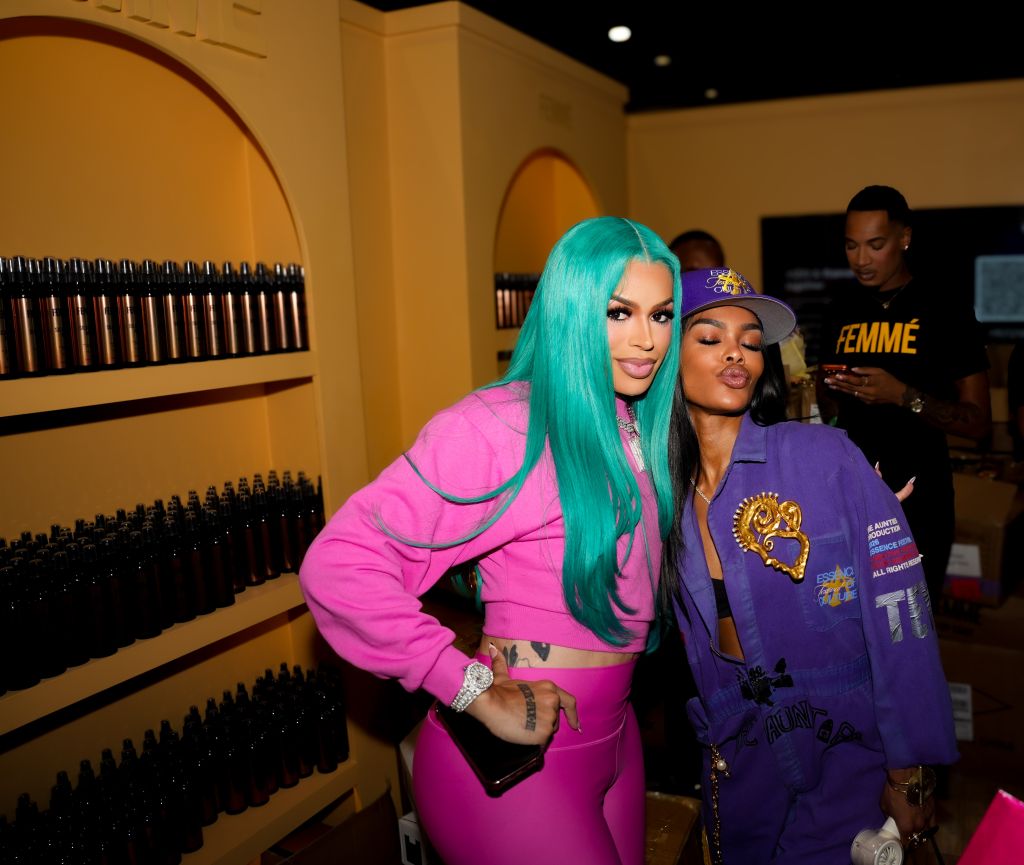 Two women with colorful outfits and hairstyles posing together in a room with shelves of bottles.
