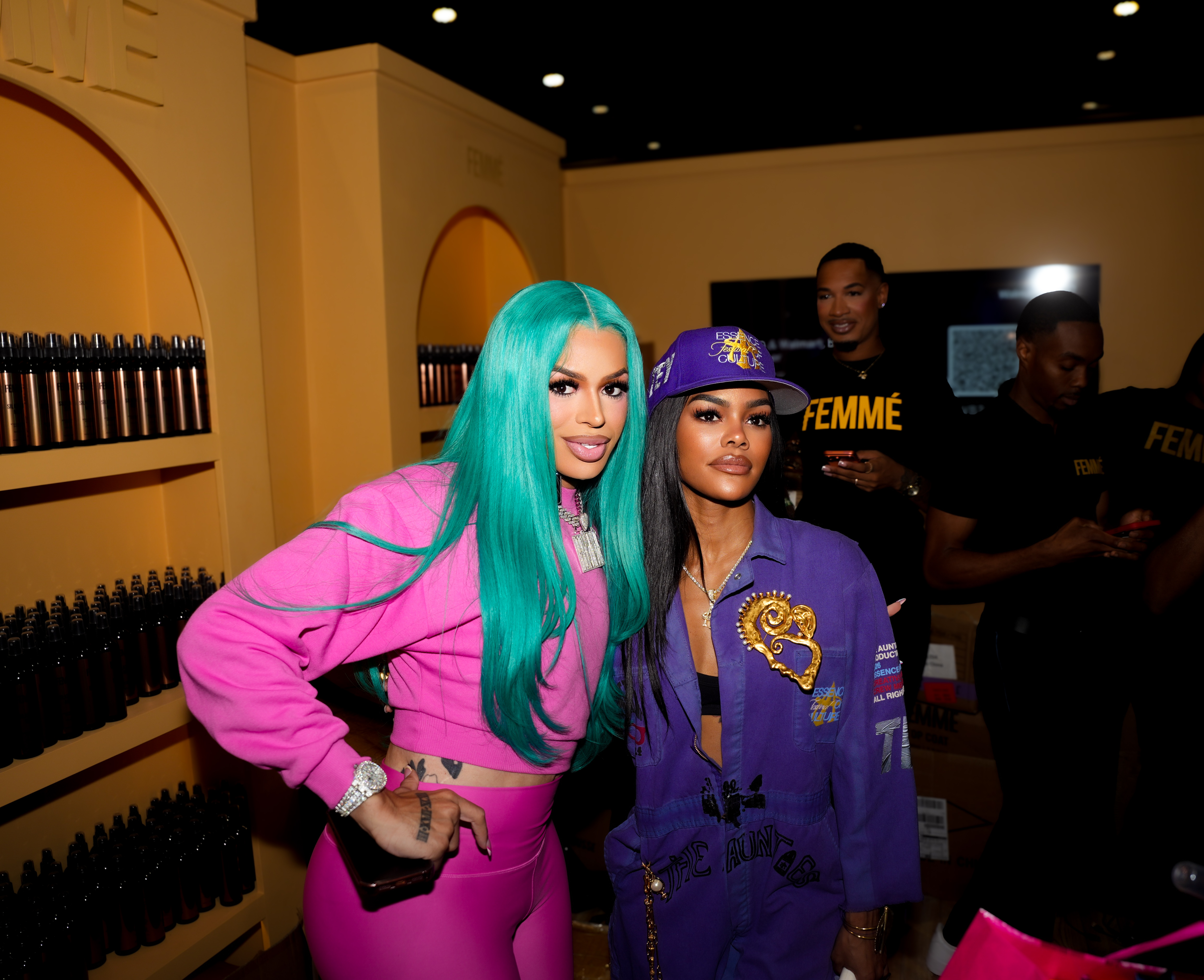 Two women wearing colorful, coordinated outfits posing together in a room with shelves of products behind them. One has teal hair and the other is wearing a purple hat with "FEMME" printed on it.