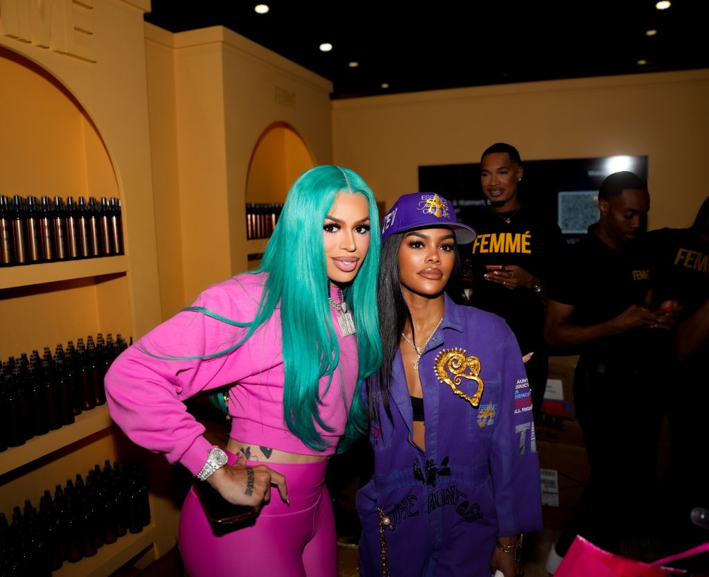 Two women wearing colorful, coordinated outfits posing together in a room with shelves of products behind them. One has teal hair and the other is wearing a purple hat with "FEMME" printed on it.