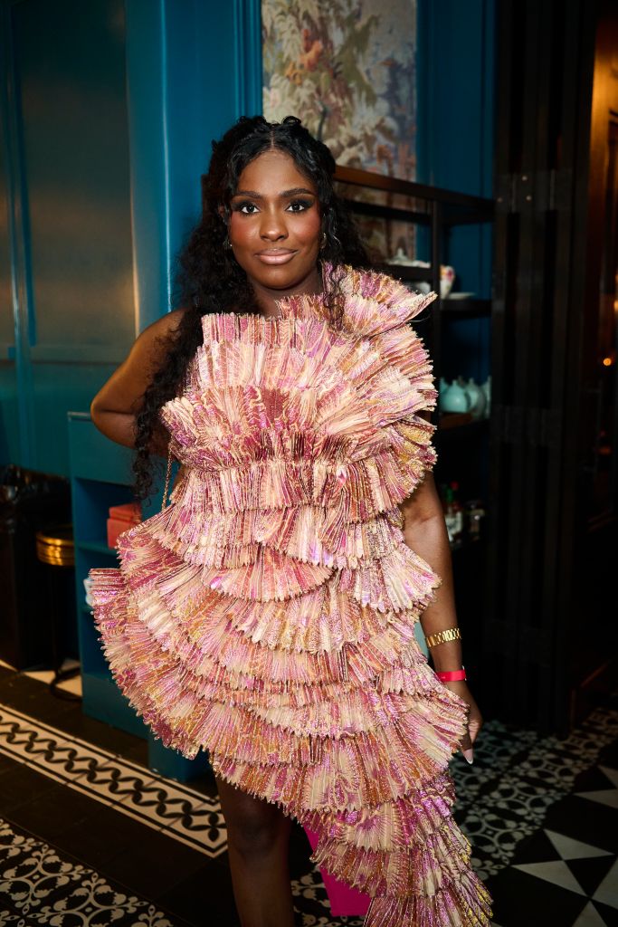 A woman wearing a vibrant, ruffled pink and gold dress stands in a room with a colorful painting on the wall behind her.
