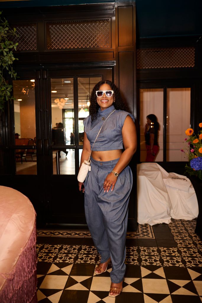 A woman in a gray crop top and pants stands in a dimly lit room with ornate wooden panels, smiling and wearing sunglasses.