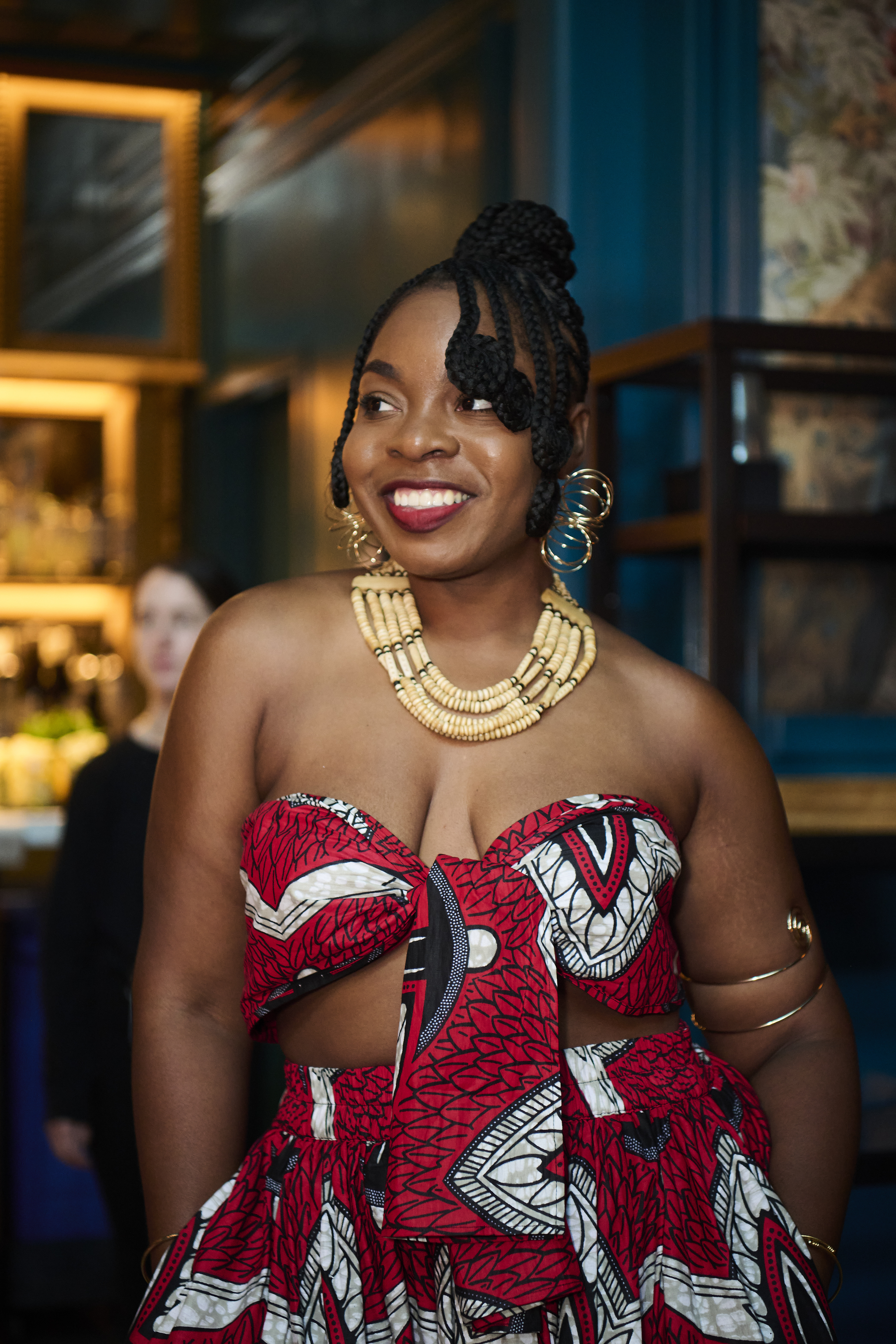 A smiling woman wearing a red and white patterned dress, large hoop earrings, and a beaded necklace stands in a dimly lit room.