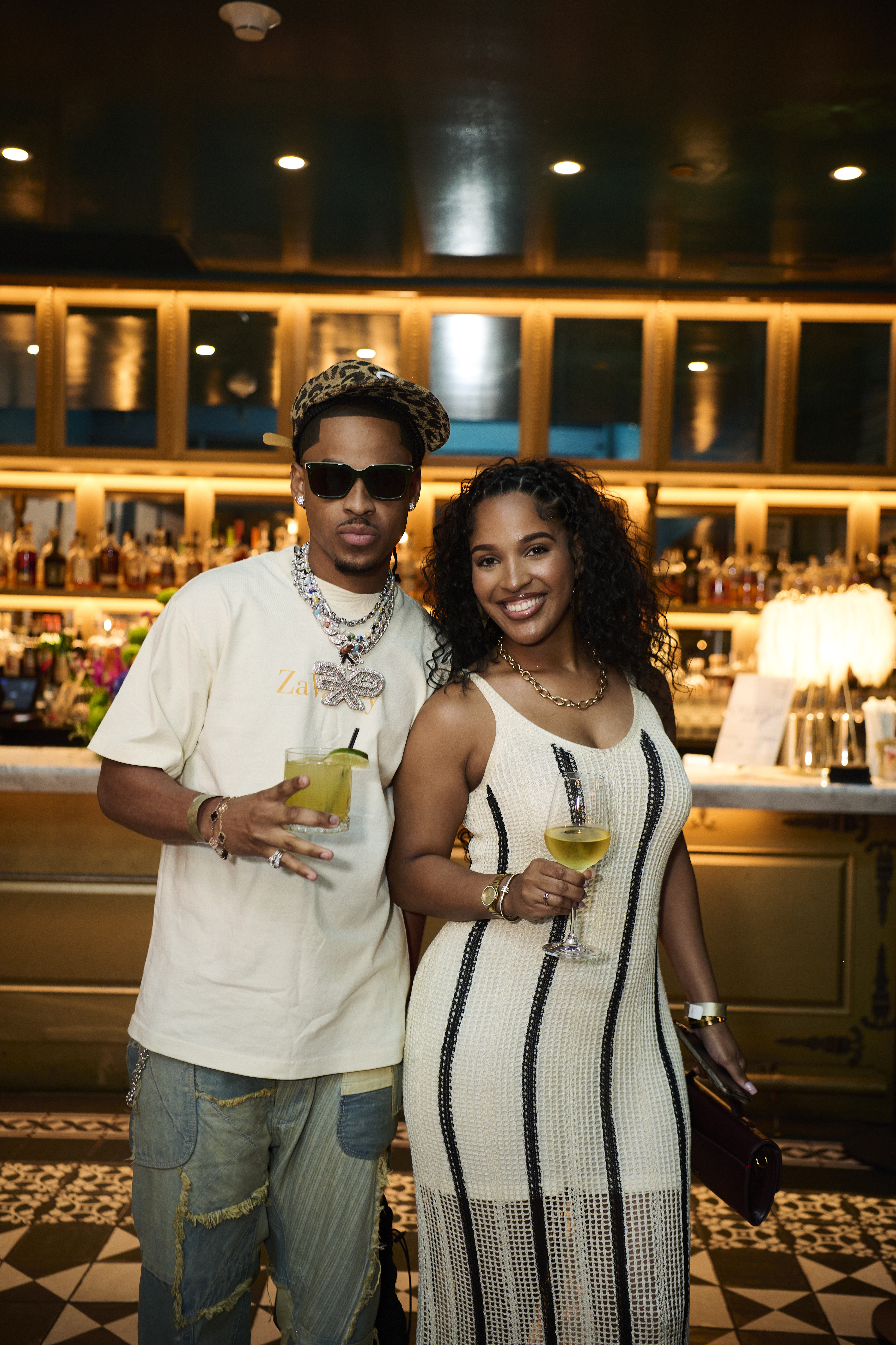 Two people, a man and a woman, standing together in a bar or restaurant setting, both holding drinks and smiling.