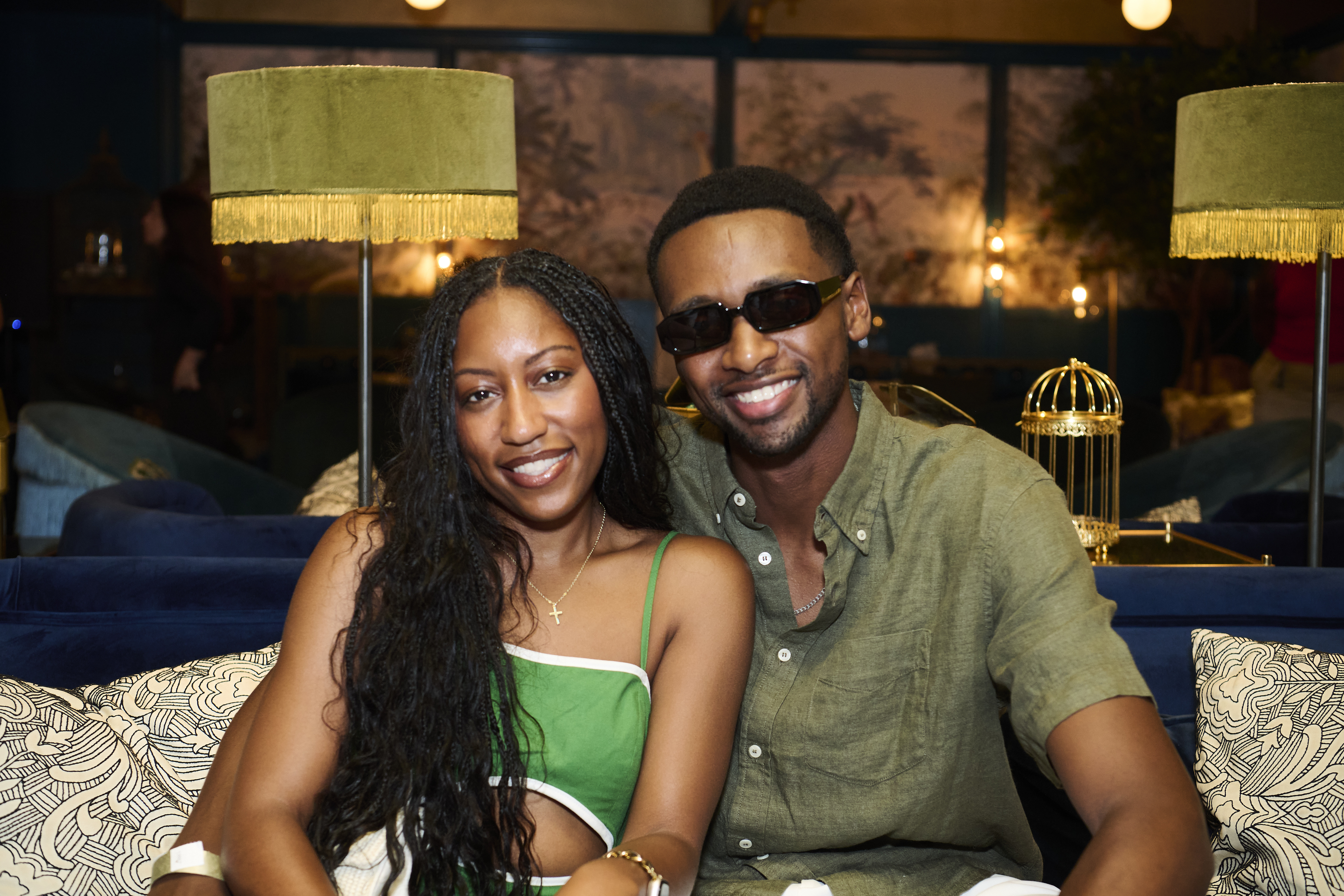 A smiling couple sitting on a couch in a dimly lit room, with lamps and decorative elements visible in the background.