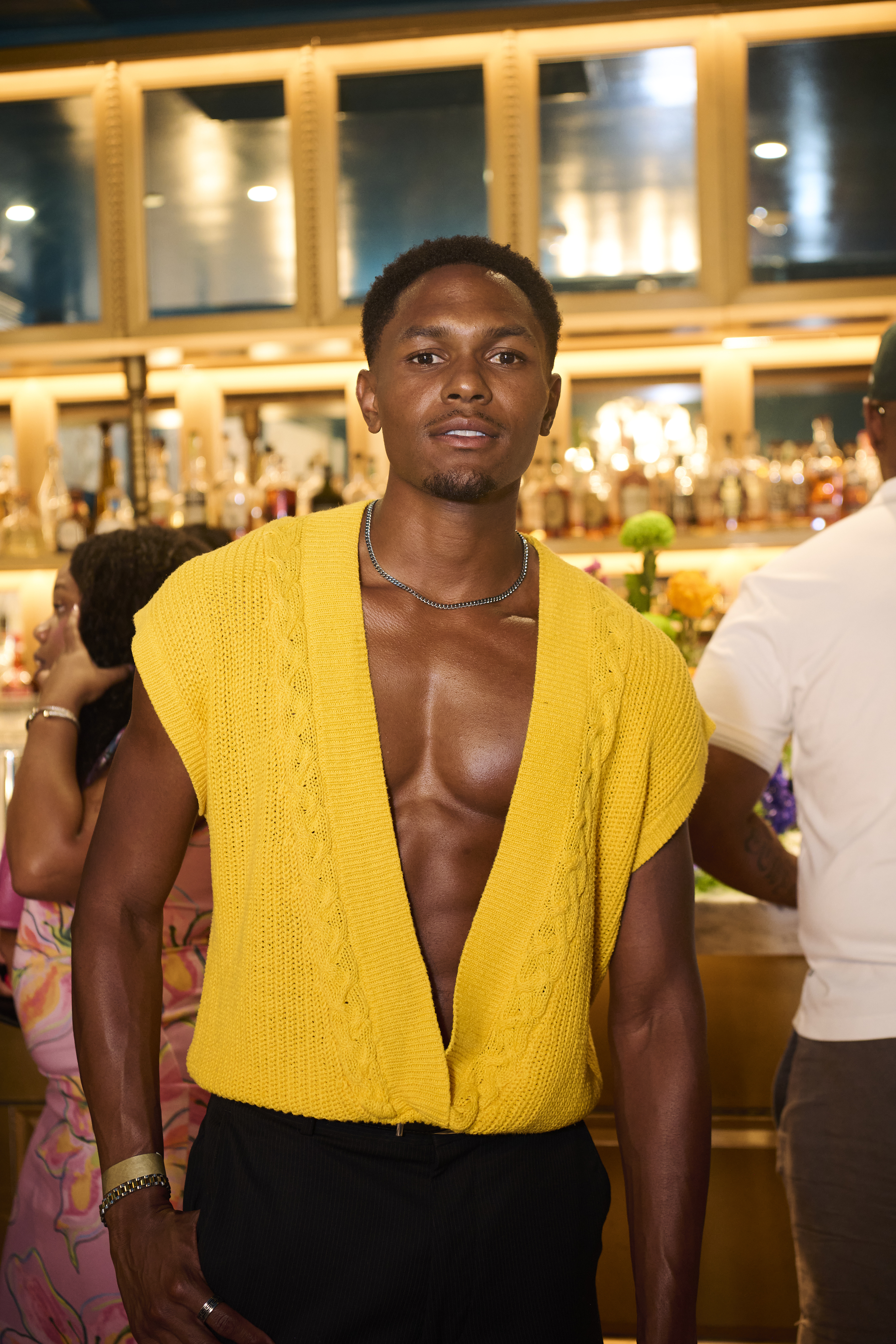 A shirtless man wearing a yellow knit cardigan stands in a dimly lit room with a bar in the background.
