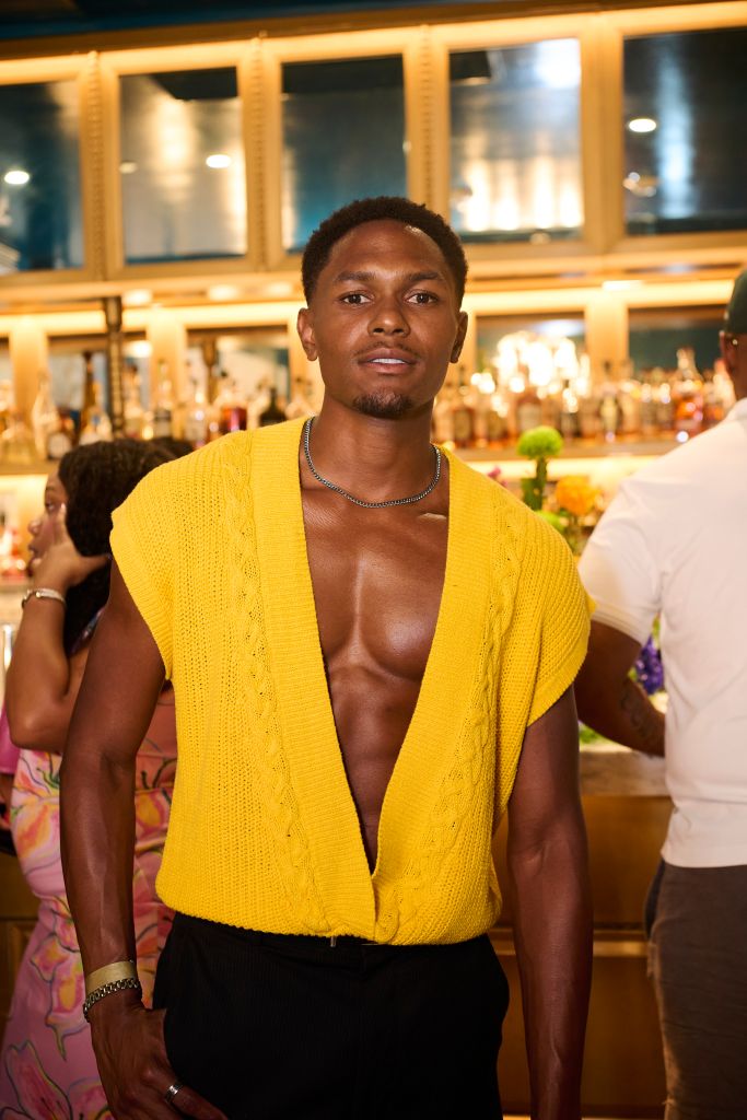 A shirtless man wearing a yellow knit cardigan stands in a dimly lit room with a bar in the background.