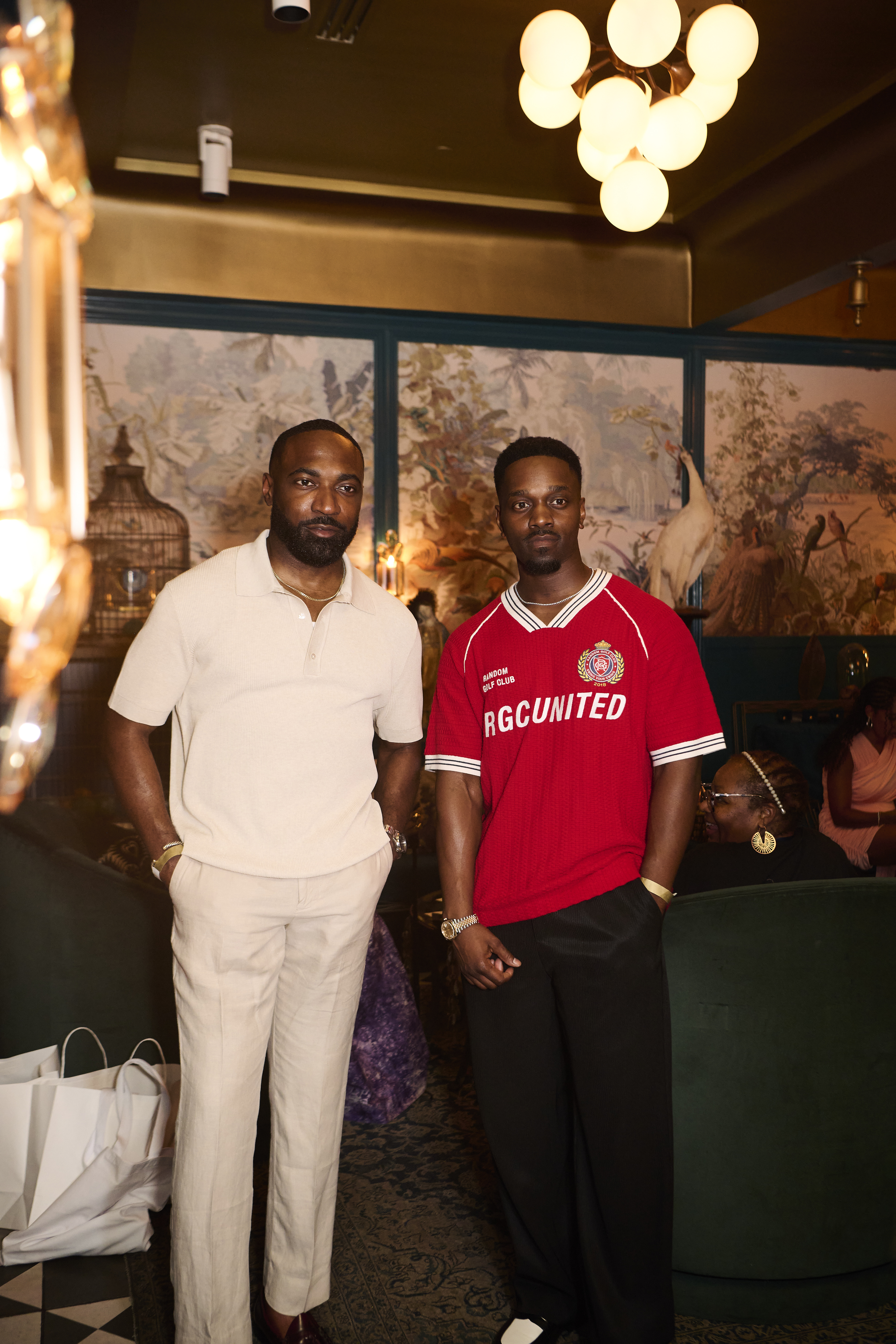 Two Black men standing in a room, one wearing a white shirt and the other wearing a red jersey with "RGUNITED" printed on it.