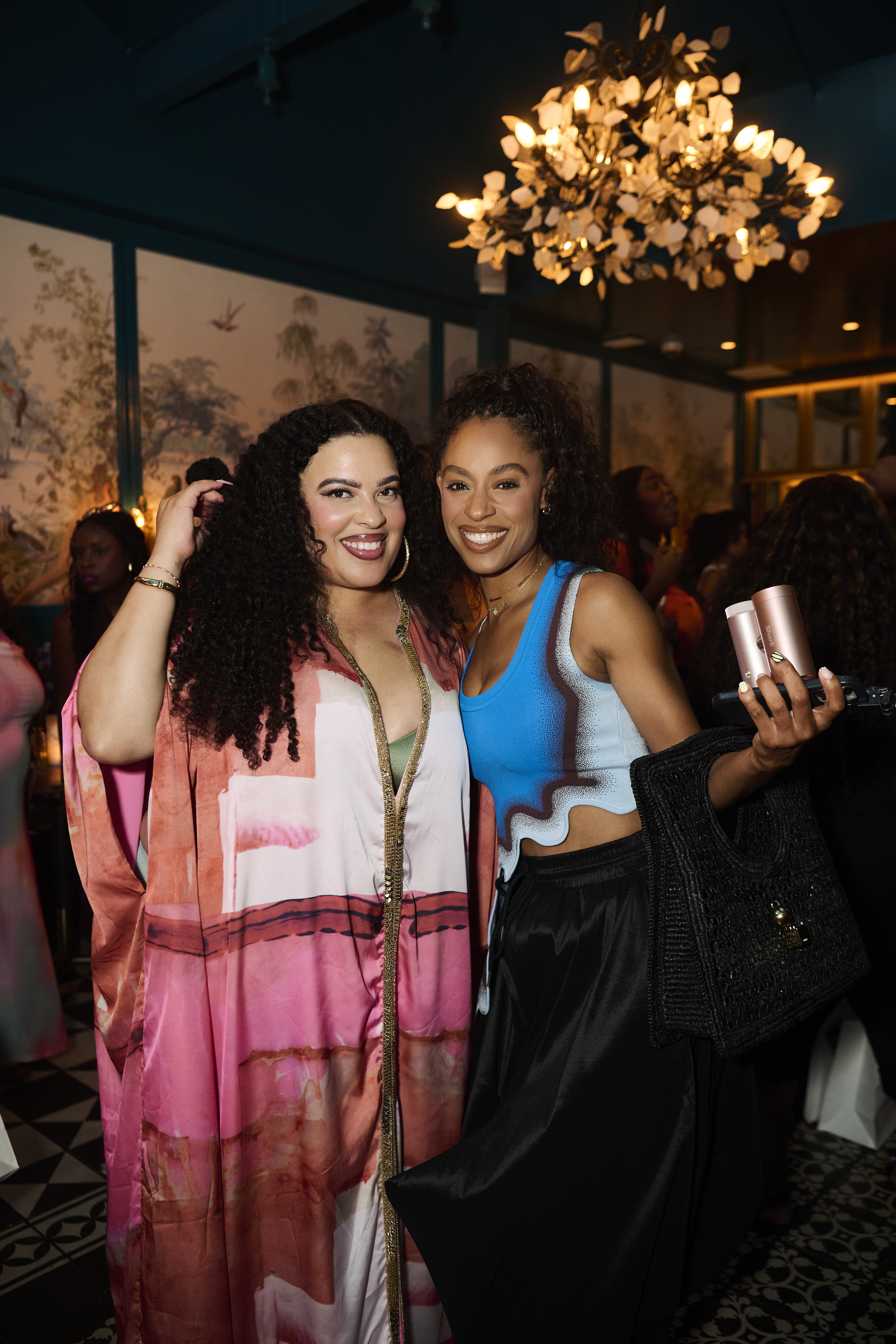 Two smiling women in colorful outfits posing for a selfie in a dimly lit room with a chandelier overhead.