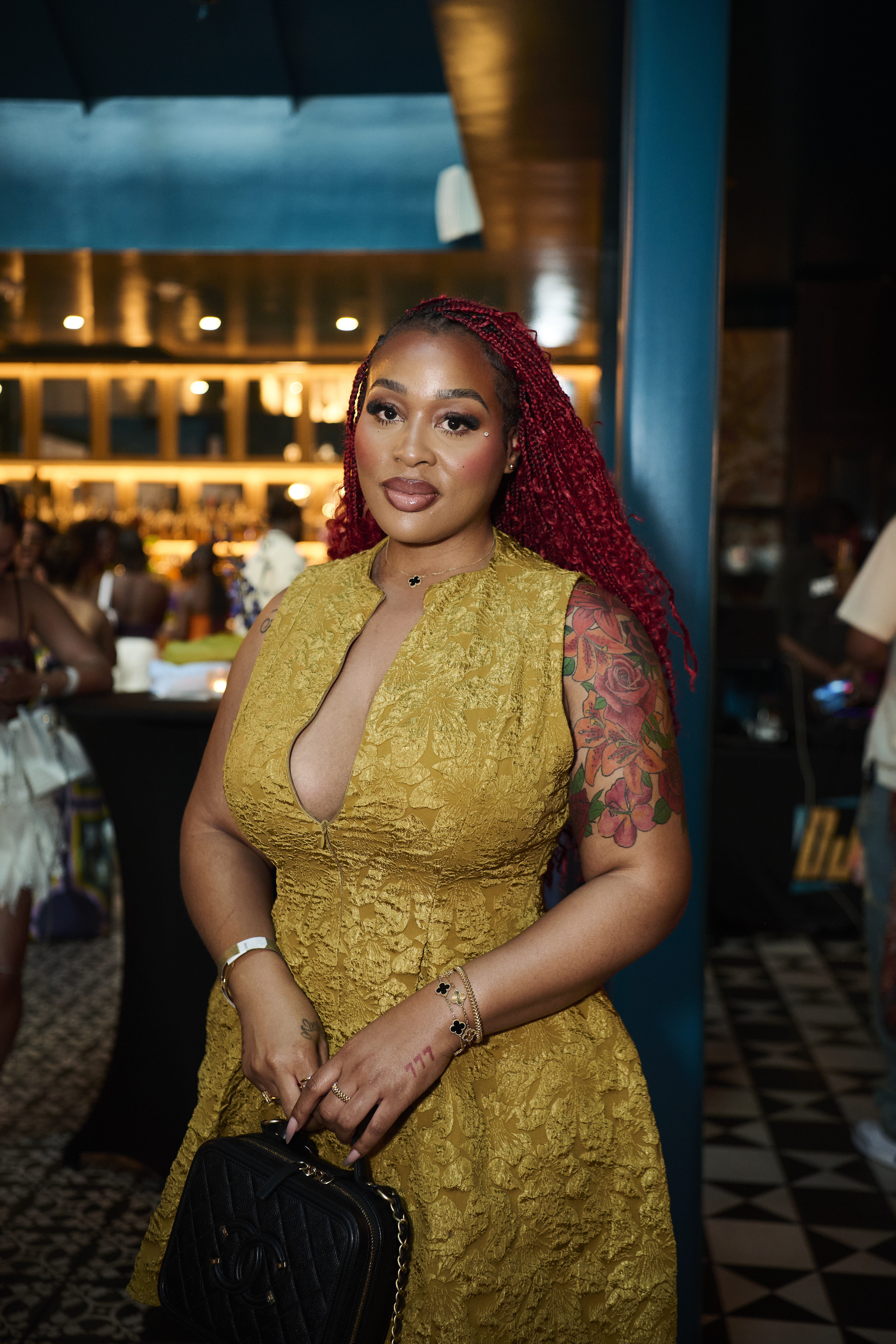 A woman with red braided hair wearing a yellow lace dress and tattoos on her arm, standing in a dimly lit setting.