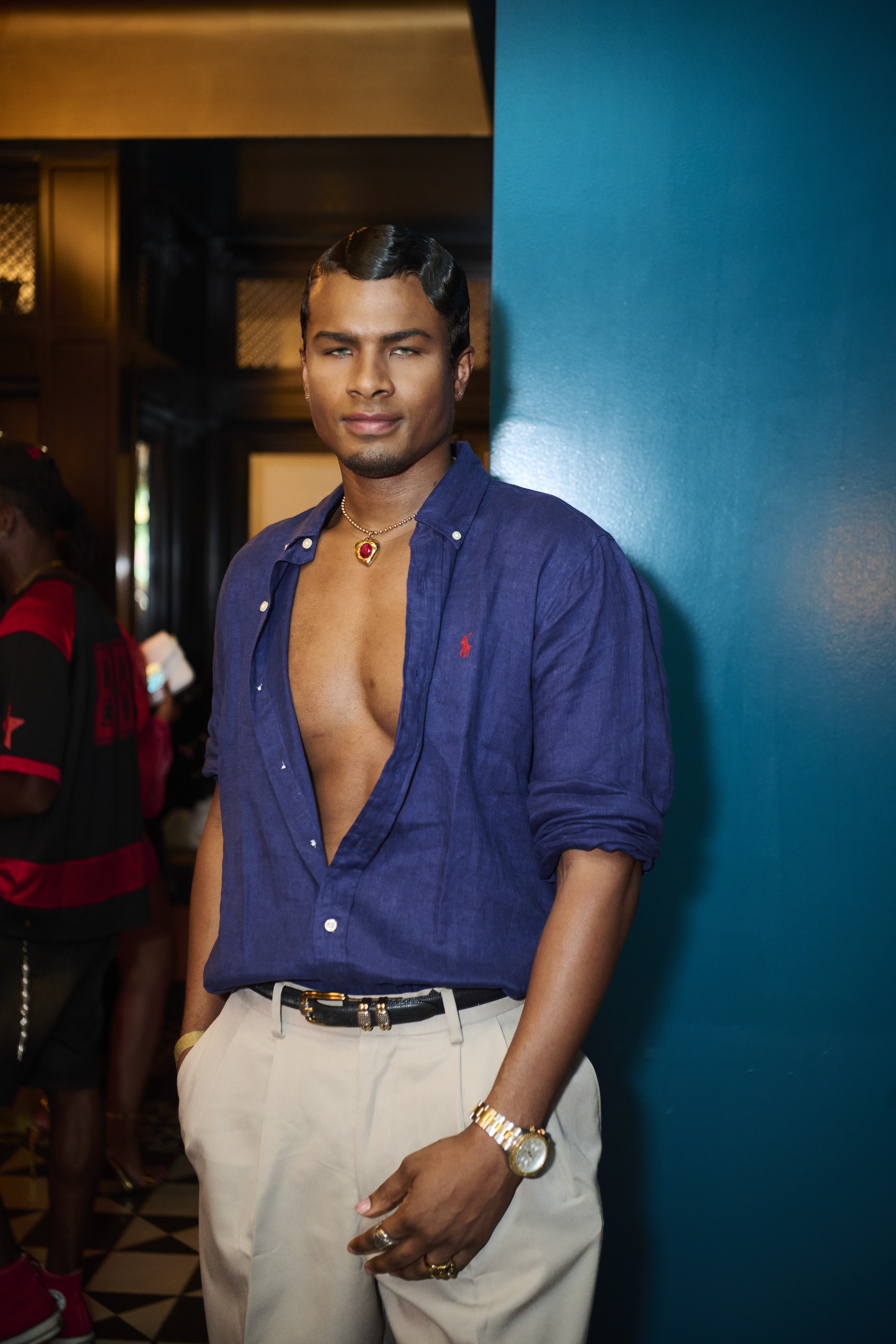 A man wearing a navy blue button-down shirt and white pants stands in front of a blue wall, with a necklace and watch visible.