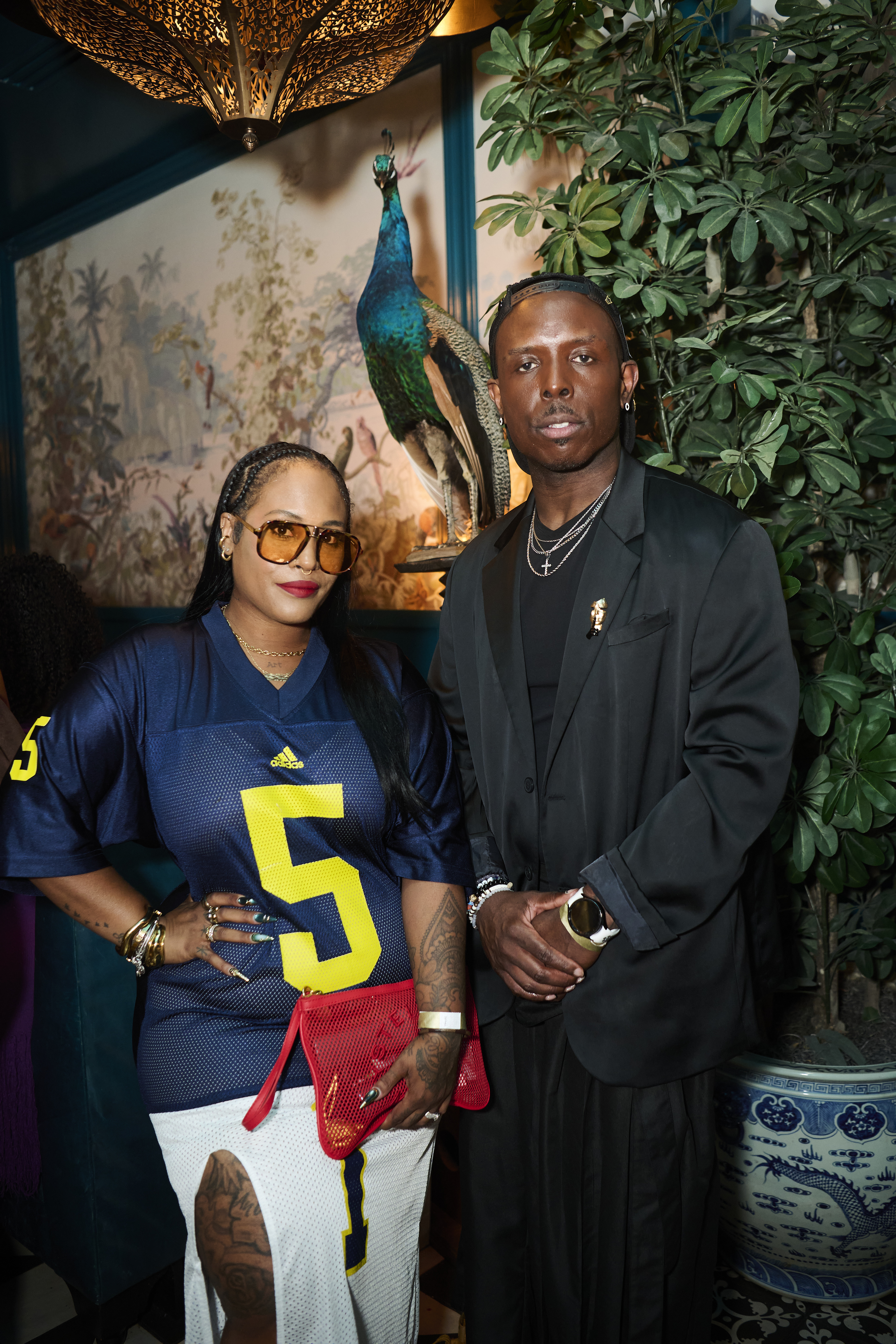 Two people, a woman wearing a blue and yellow sports jersey and a man in a black suit, standing in front of a tropical plant and artwork with a peacock.