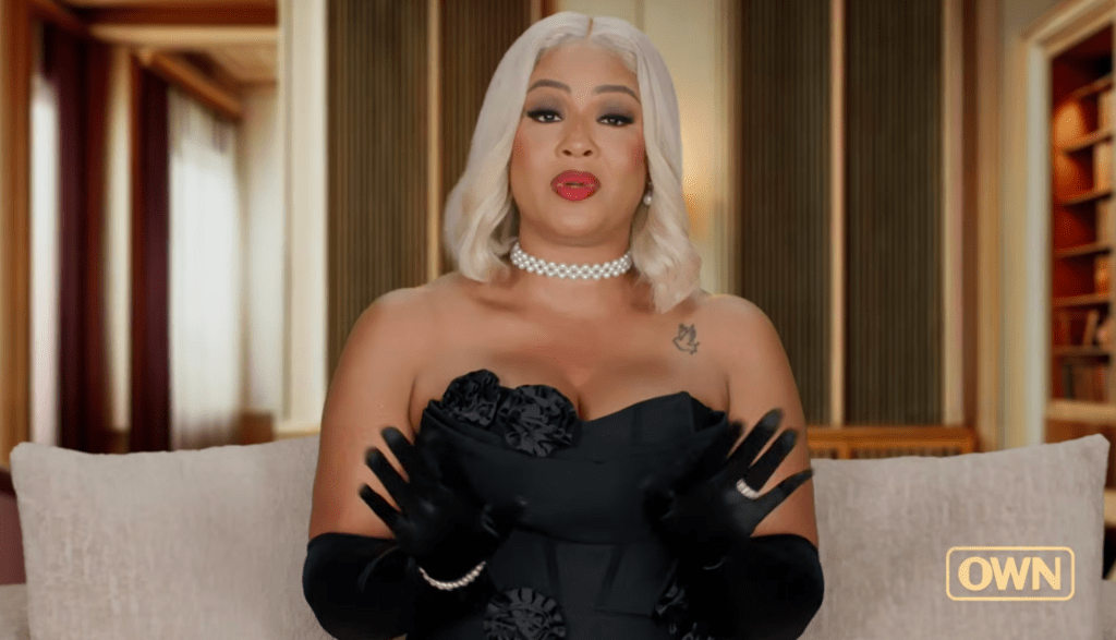 A person with long white hair and bold makeup wearing a black strapless dress and gloves, posing in a room with wooden paneling.