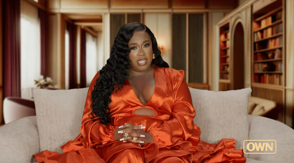 A woman in an orange satin dress sitting on a couch, with a serious expression on her face and her hands clasped in front of her.