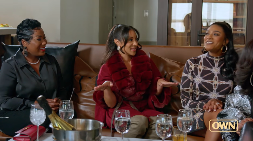 Three women sitting on a couch, one wearing a red coat, engaged in conversation and laughter.
