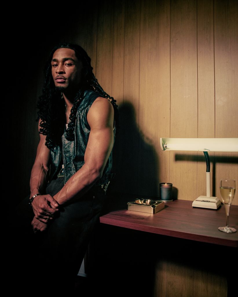A man with dreadlocks wearing a green sleeveless vest stands in a dimly lit room, with a table in front of him containing a candle, glass, and other objects.