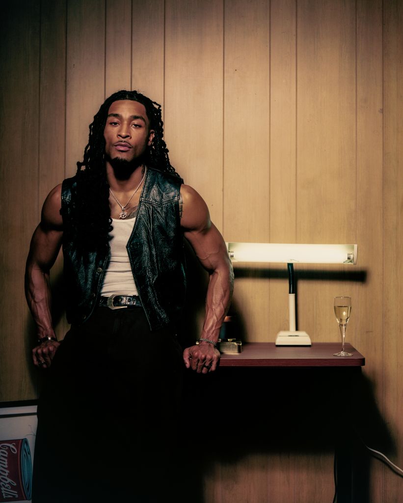 A muscular man with dreadlocks wearing a leather jacket stands in front of a wooden wall, with a glass of champagne on a table beside him.