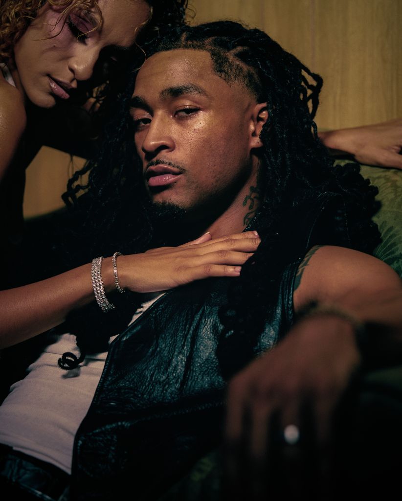 A close-up portrait of a man with dreadlocks, wearing a dark shirt, looking pensive and introspective.
