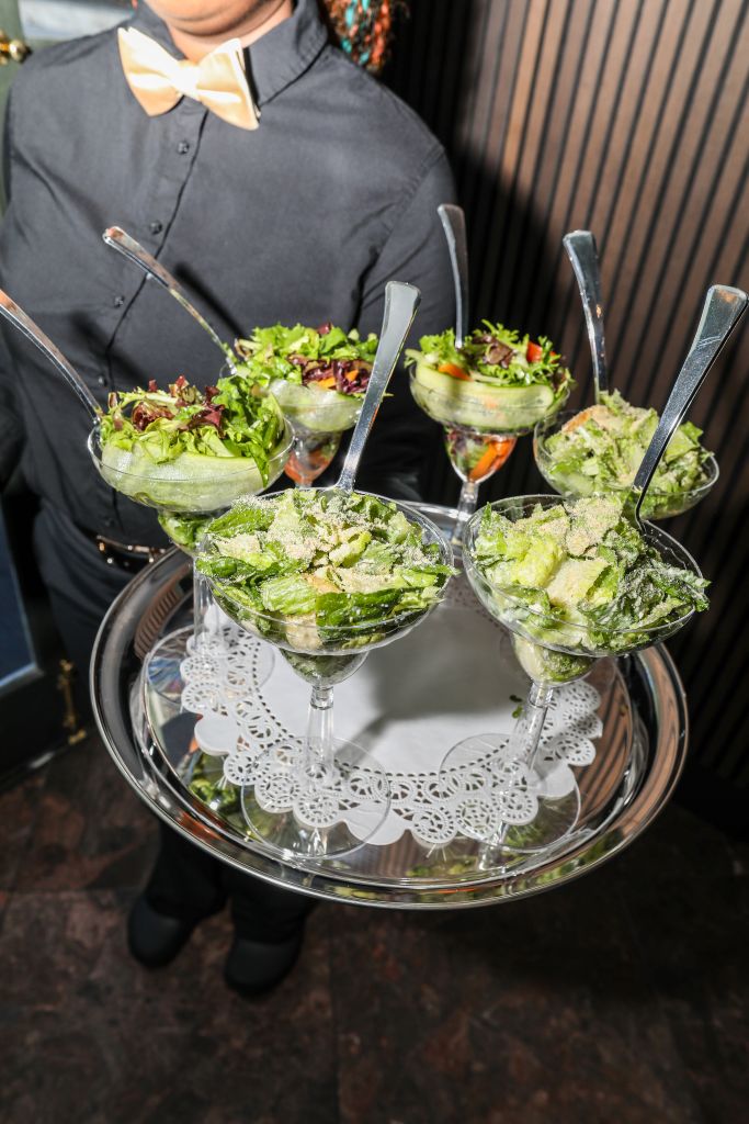 A server in a black uniform holding several glasses filled with a green salad or lettuce mixture on a tray with lace doilies.