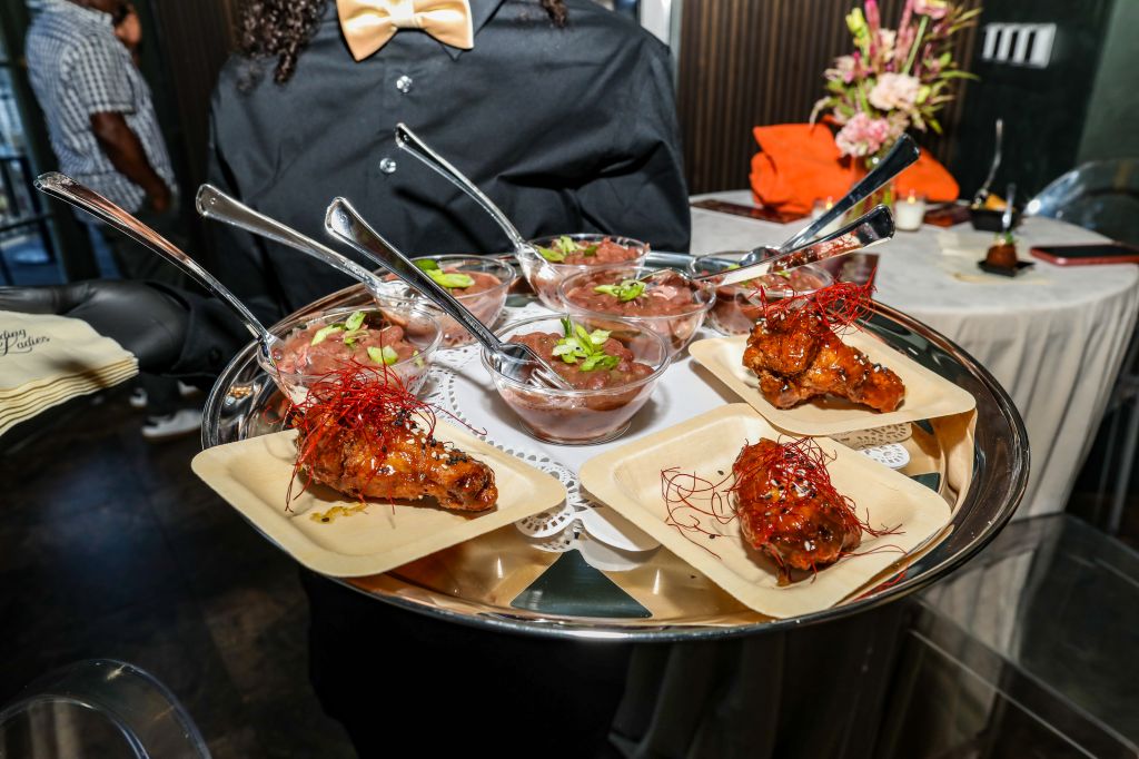 A tray of appetizers featuring glazed chicken wings and small bowls of what appears to be a tomato-based dipping sauce, garnished with fresh herbs, on a table with a floral arrangement.