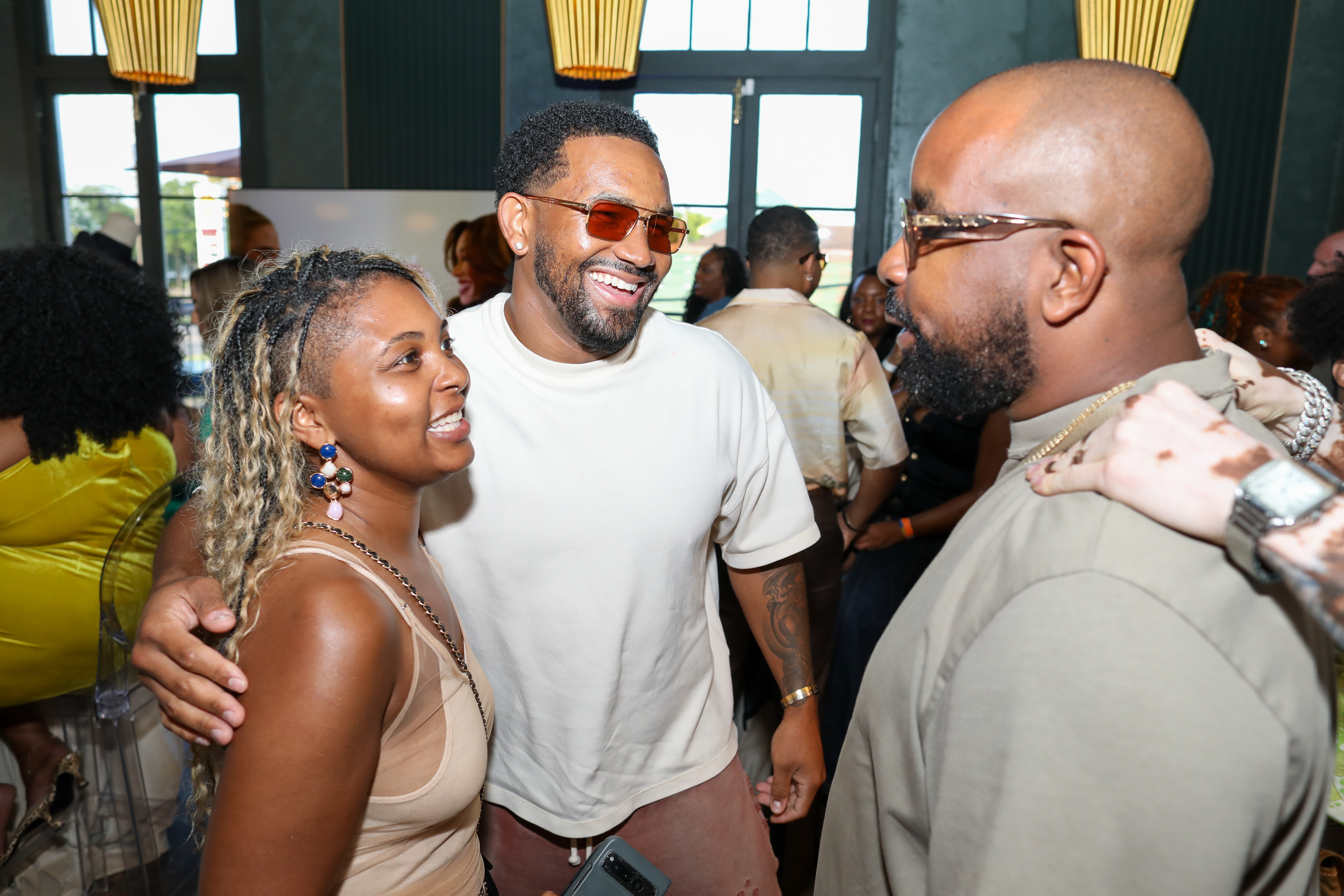 Three people smiling and embracing at an event, with one person wearing sunglasses.