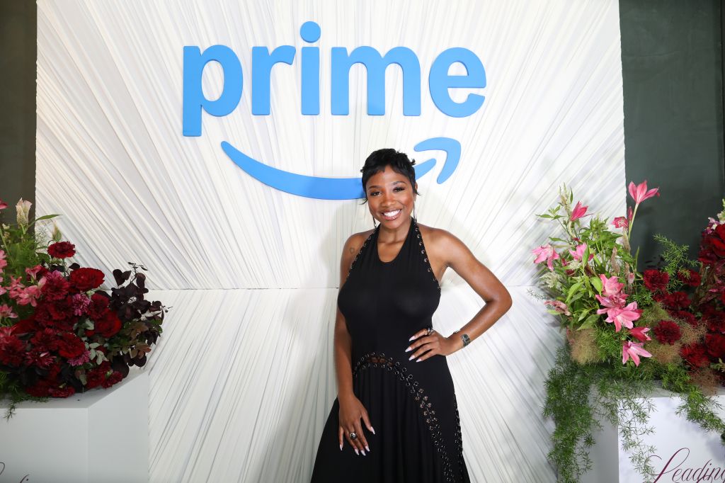 A smiling woman in a black dress standing in front of a large "Prime" logo backdrop with floral arrangements.