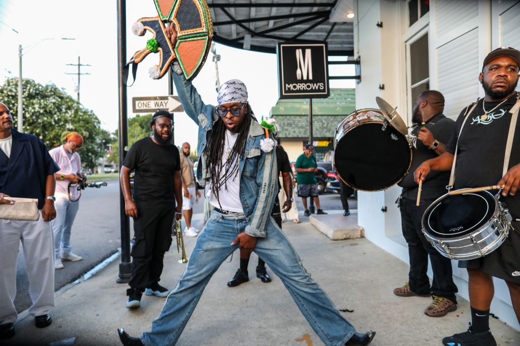 A group of people, including a person wearing a dreadlock wig and holding a drum, standing outside a building with a "Morrows" sign.