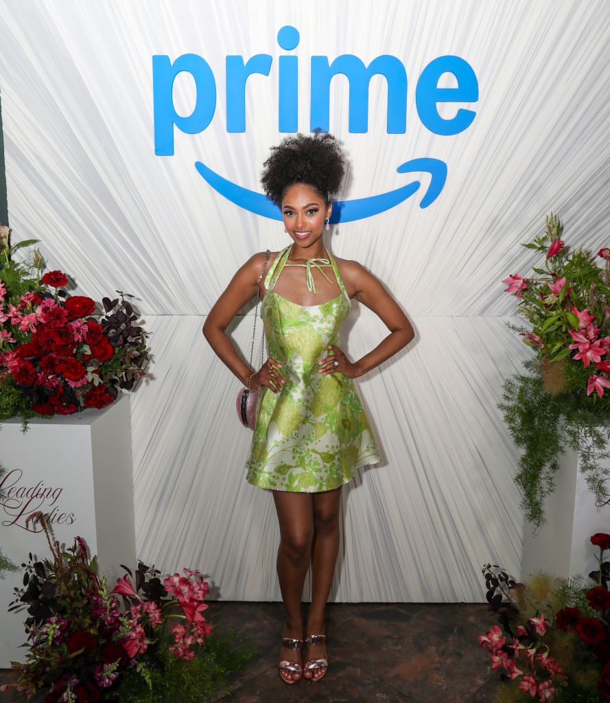 A woman in a green floral dress standing in front of a large "Prime" logo backdrop, surrounded by floral arrangements.