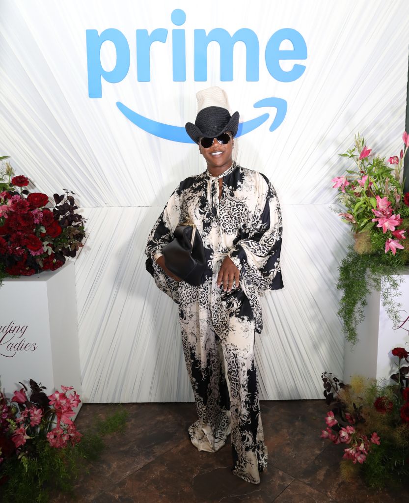 A person wearing a black and white patterned outfit and sunglasses stands in front of a backdrop with the Amazon Prime logo.
