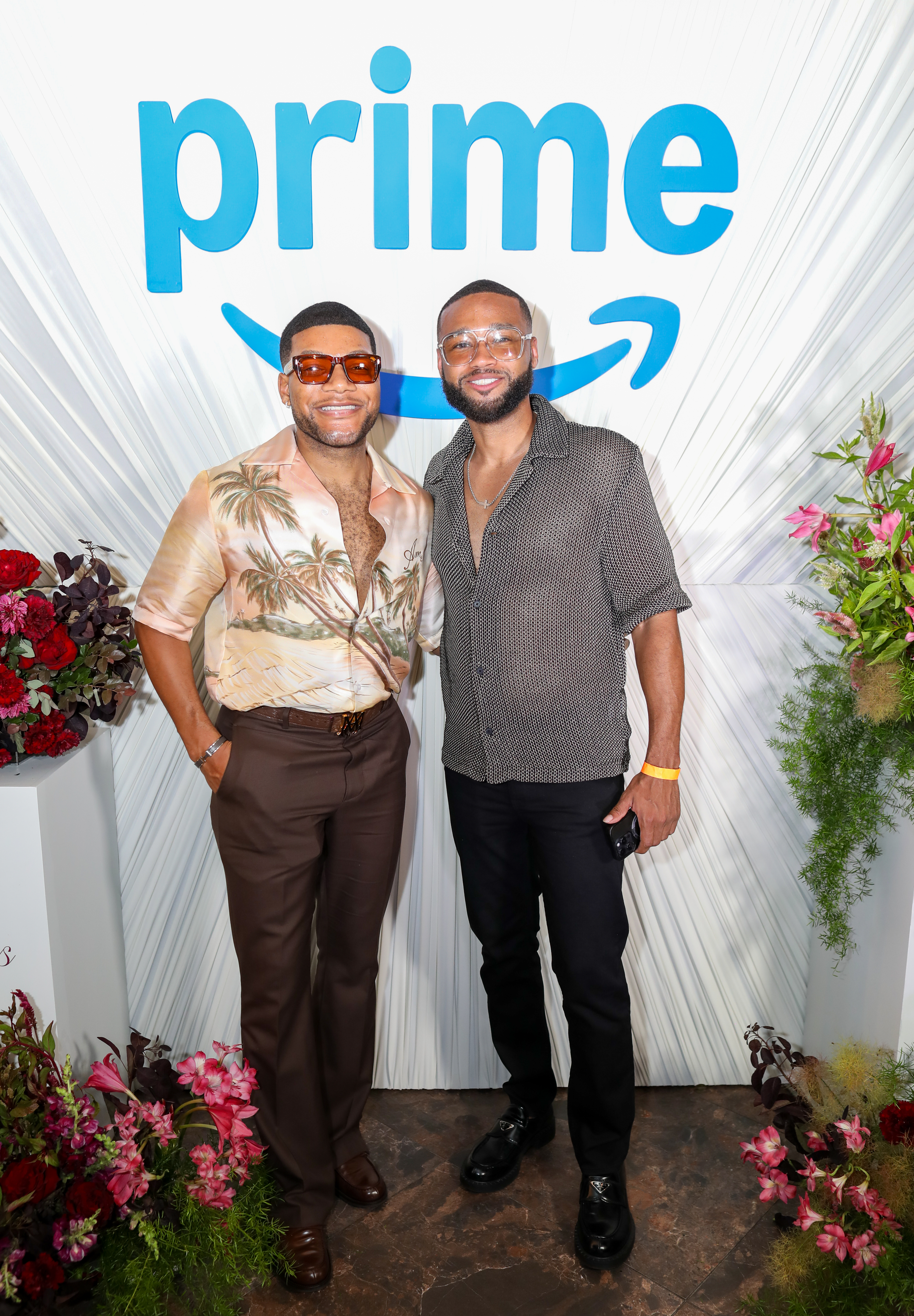 Two men posing in front of a "Prime" backdrop, one wearing a tropical print shirt and the other a patterned button-up.