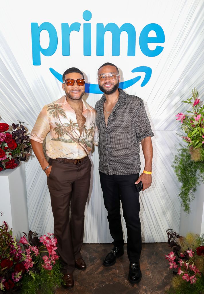 Two men posing in front of a "Prime" backdrop, one wearing a tropical print shirt and the other a patterned button-up.