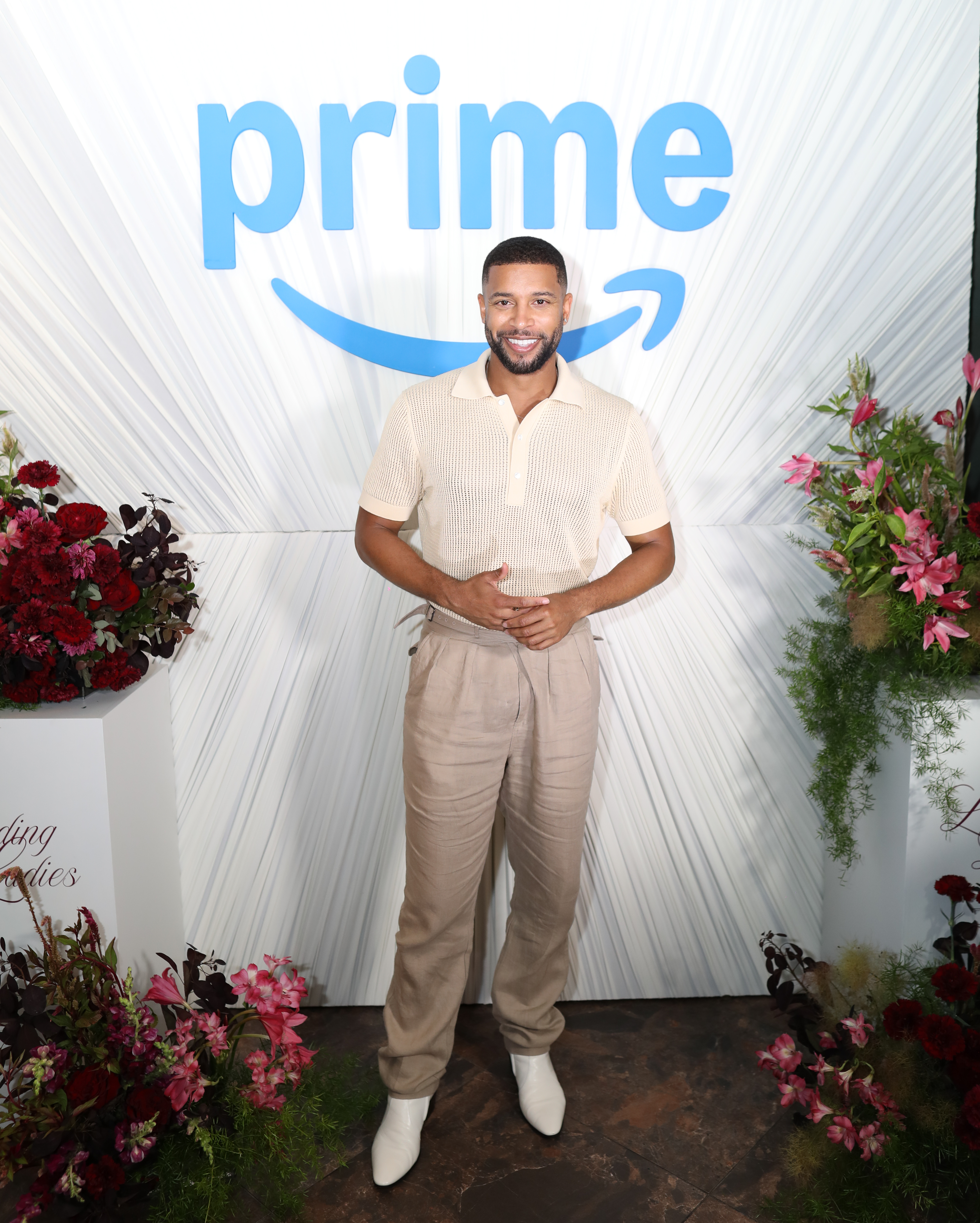 A smiling man wearing a beige outfit stands in front of a large "Prime" logo backdrop, surrounded by floral arrangements.