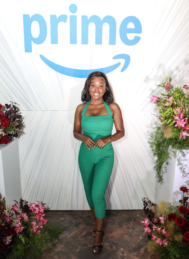 A smiling woman in a green jumpsuit standing in front of a large Amazon Prime logo backdrop, surrounded by floral arrangements.
